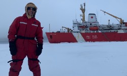 A person in red coast guard winter clothing standing on a plain of ice in front of the coast guard vessel