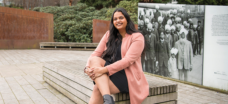 A student with long, dark hair sits in front of a large photo showing Sikh people in 1914.