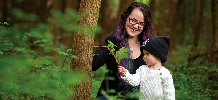 A woman smiles lovingly at a toddler in a hat who is looking curiously at a leaf in a wooded area.
