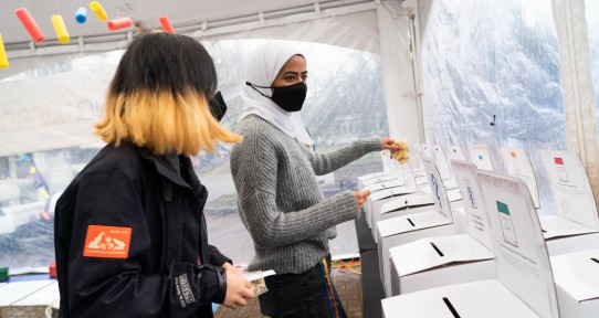 Two students stand facing several white boxes with slots in them