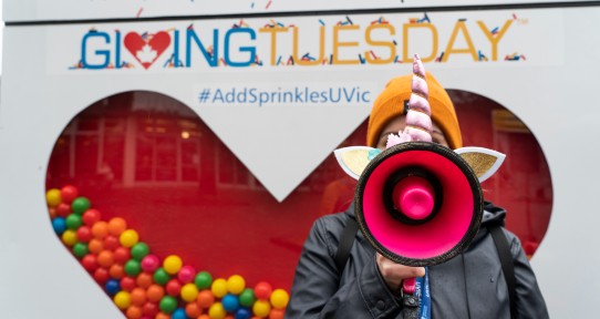 A women holding a unicorn shaped megaphone to her mouth stands in front of a display of a heart, half full of colourful balls