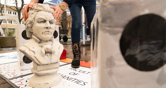 A pair of hands lift a replica of a bust of a man's head. In the foreground is a large die.