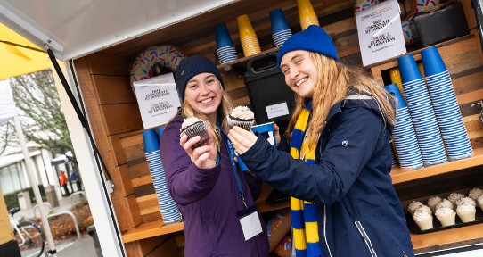 Two female students wearing hats and scarves hold cupcakes