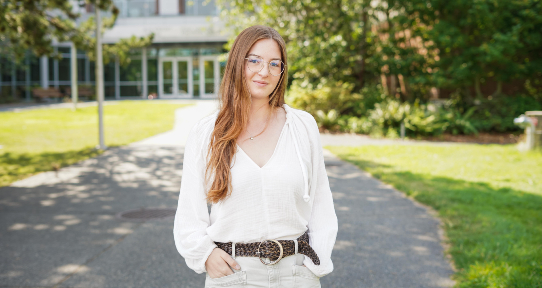 A student with long hair and glasses in a white blouse stands on the path leading to the UVic Bob Wright building