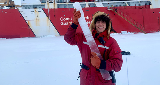 Man in red survival suit holding column on ice in front of coast guard vessel