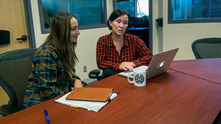 Two people seated at a table talking about what they see on a laptop