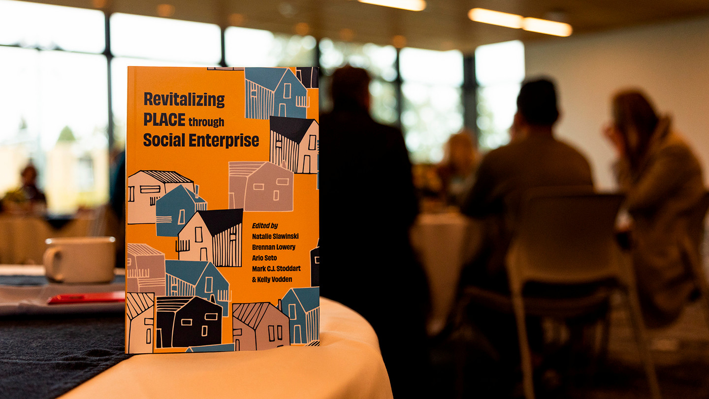 A book called Revitalizing PLACE through Social Enterprises sits on a table at a book launch with people in the background