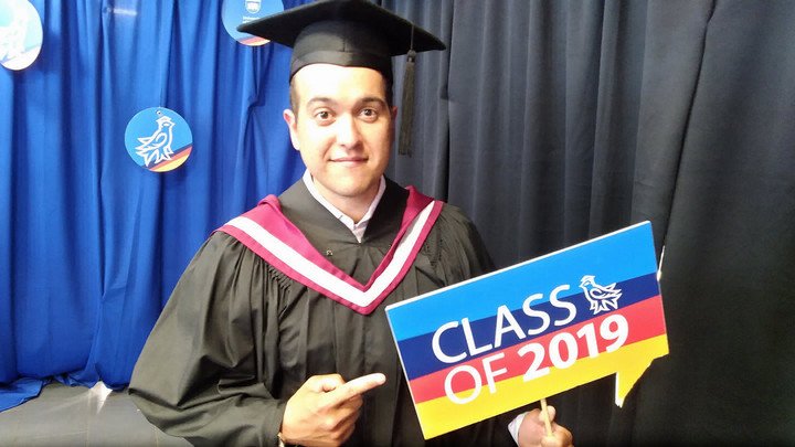 Felipe Ferreira at this graduation ceremony with the cap and gown pointing to a Class of 2019 sign