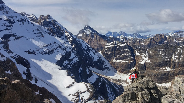 Felipe Ferreira at a top of a snowy mountain peak clutching a Canada flag with more snowy mountain peaks in the distance