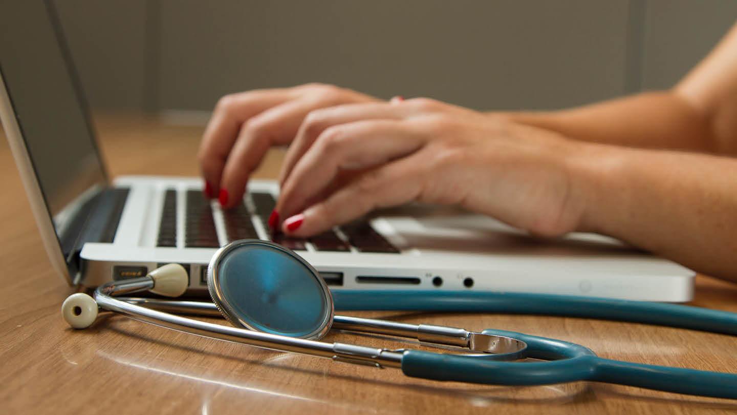 Person sitting while using laptop computer with a green stethoscope nearby