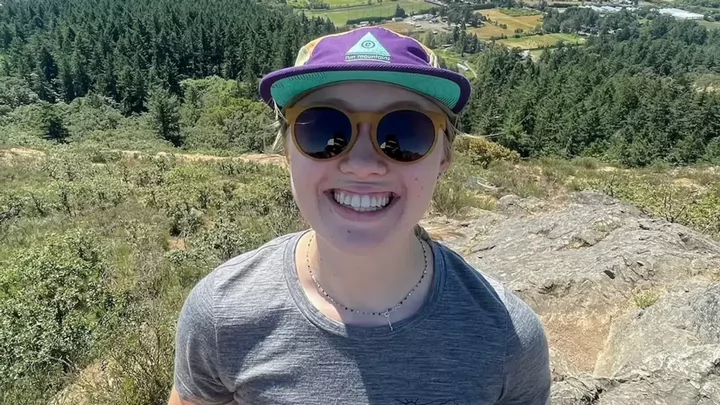 Person with hat and sunglasses standing on top of a hill, smiling in closeup 