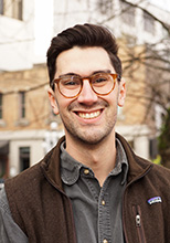 Aaron Bailey, PhD student in SDH. Aaron has short dark brown hair that is slicked back at the front. He wears round tortoise shell coloured glasses and a brown zip-up Patagonia vest over a medium gray button-up shirt. He stands in front of the Cornett Building on UVic campus.