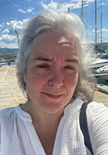 Andrea White, PhD student in SDH. Andrea has light grey hair. She is wearing a white linen shirt with a V-neck. She takes a selfie on a cobblestone pier. The sky is bright blue with some clouds. Sailboats are docked on the right behind her with azure ocean on the left..