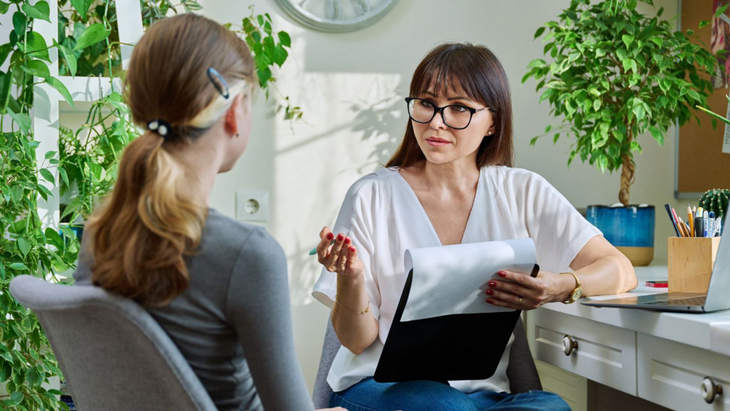 A woman with glasses and a notebook sits at a desk, engaging attentively with another woman in an office filled with lush green plants.