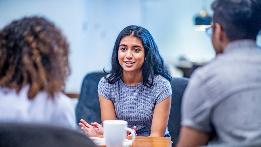 A woman with dark hair is speaking and gesturing with her hands in a casual setting, while two others listen attentively.