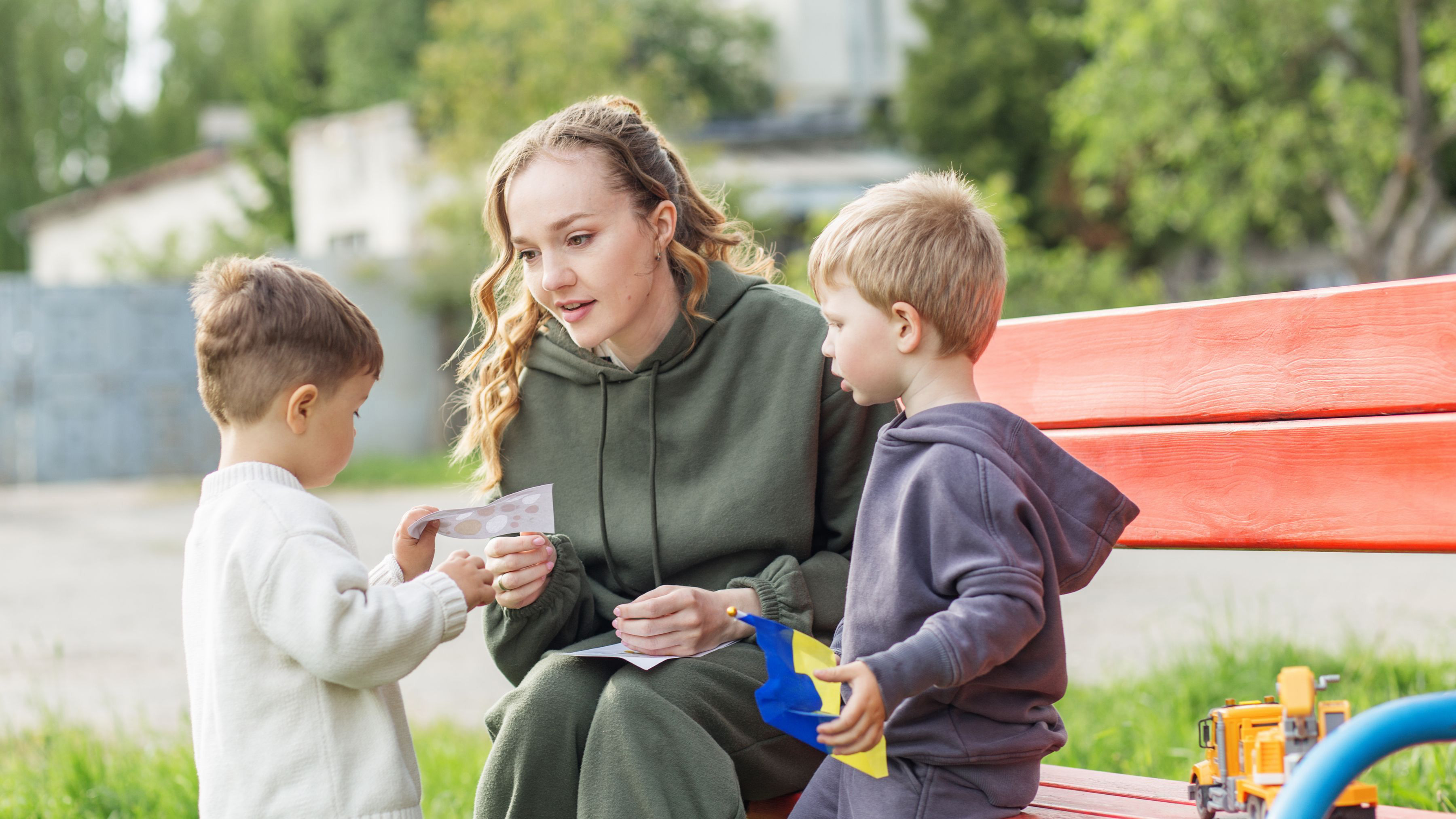 A woman in a green hoodie sits on a red bench, engaging with two young boys. One boy holds toy trucks while the woman examines a card. The scene is outdoors, surrounded by greenery.