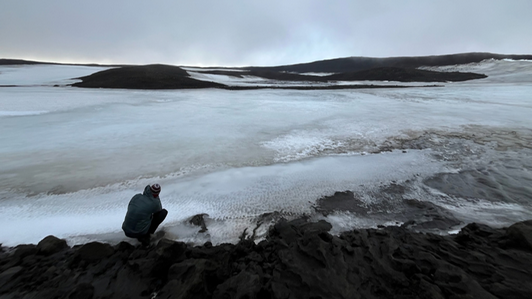 Braedon Lowey kneels at the edge of a glacier, filming.