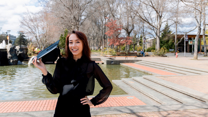 Andini Makosinksi, a young UVic graduate, is posing in front of the Petch fountain, holding a graduation cap and smiling. It's a fall day with a blue sky.