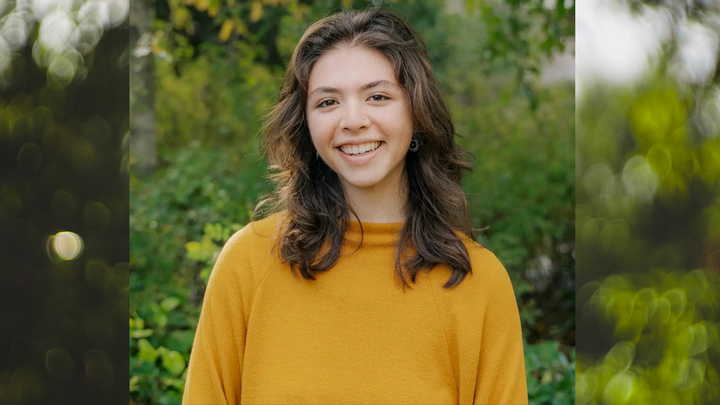 Ana Galicia wearing a yellow sweater in front of a blurry green background suggesting trees.