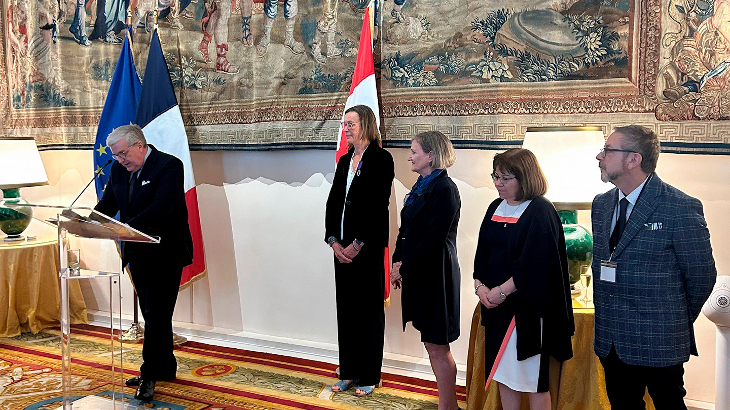 The French Ambassador to Canada is at a clear podium in an ornately decorated room with French and Canadian flags. Catherine Caws stands behind and to the right, as well as three other award recipients.