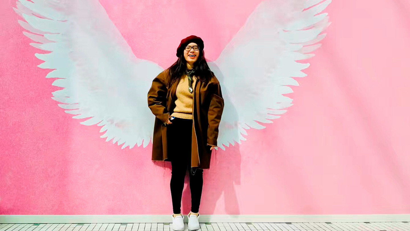 Doris Zhu poses happily in front of a pink wall, painted with white feathery angel wings.