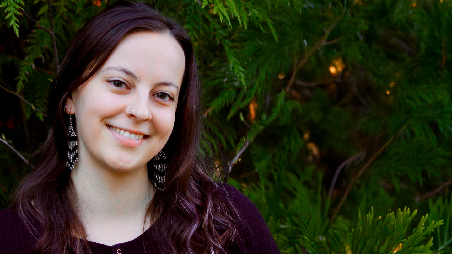 Stephanie Erickson, a Red River Metis PhD student with dark red hair, in front of background of cedar trees. She is wearing a burgundy shirt with small buttons and dangly beaded earrings.