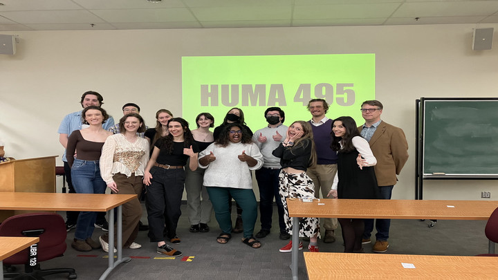 A group of students with their professor, posing in front of a projector screen that says HUMA 495.