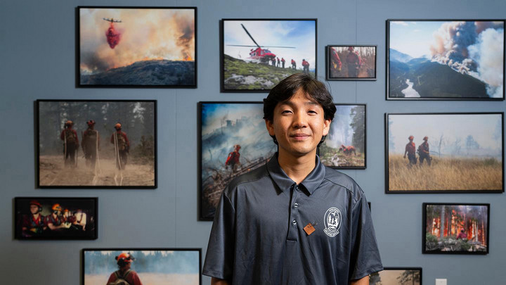 Jay Choi, a South Korean Humanities co-op student, is wearing a grey BC Wildfire Service polo shirt with a Moose Hide Campaign pin, standing in front of a wall covered in photos of firefighters combating wildfires.
