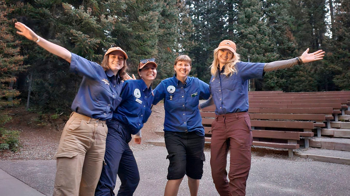 Three people in BC Parks uniforms are celebrating and smiling for the camera in front of the trees.