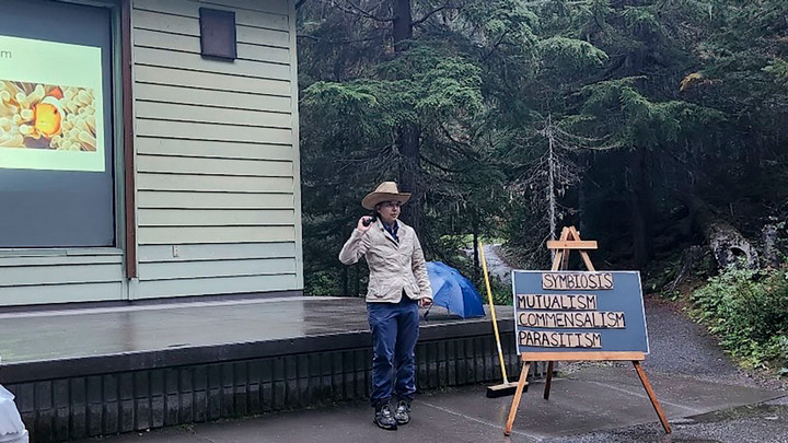 Rebecca is outside at the amphitheatre beside a sign that says "Symbiosis, Mutualism, Commensalism, Parasitism."