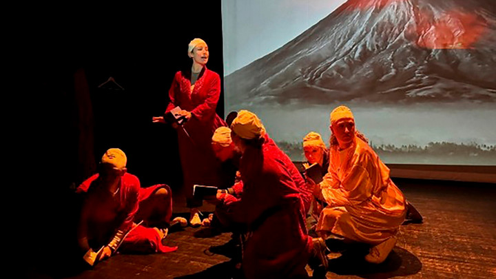 Students in red robes perform in a dimly-lit theatre with a mountain projected on a screen behind them.