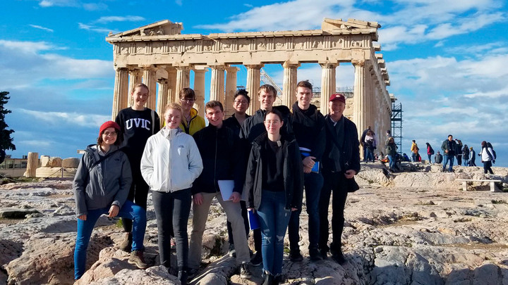 A group of UVic students pose in front of Greek ruins on a sunny, windy day. 