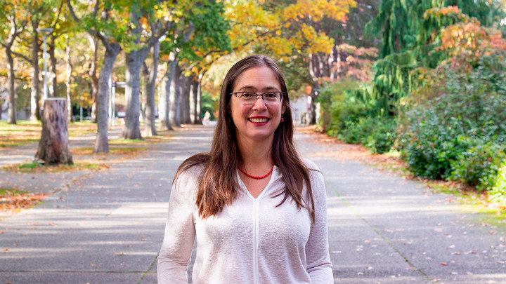 Rachel Cleves, a UVic historian with long hair and glasses, stands outside on a tree-lined walkway on UVic campus, wearing a white blouse with red lipstick.