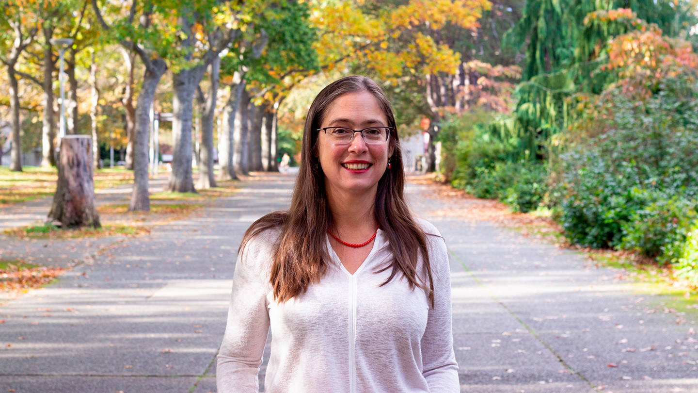Rachel Cleves, a UVic historian with long hair and glasses, stands outside on a tree-lined walkway on UVic campus, wearing a white blouse with red lipstick.