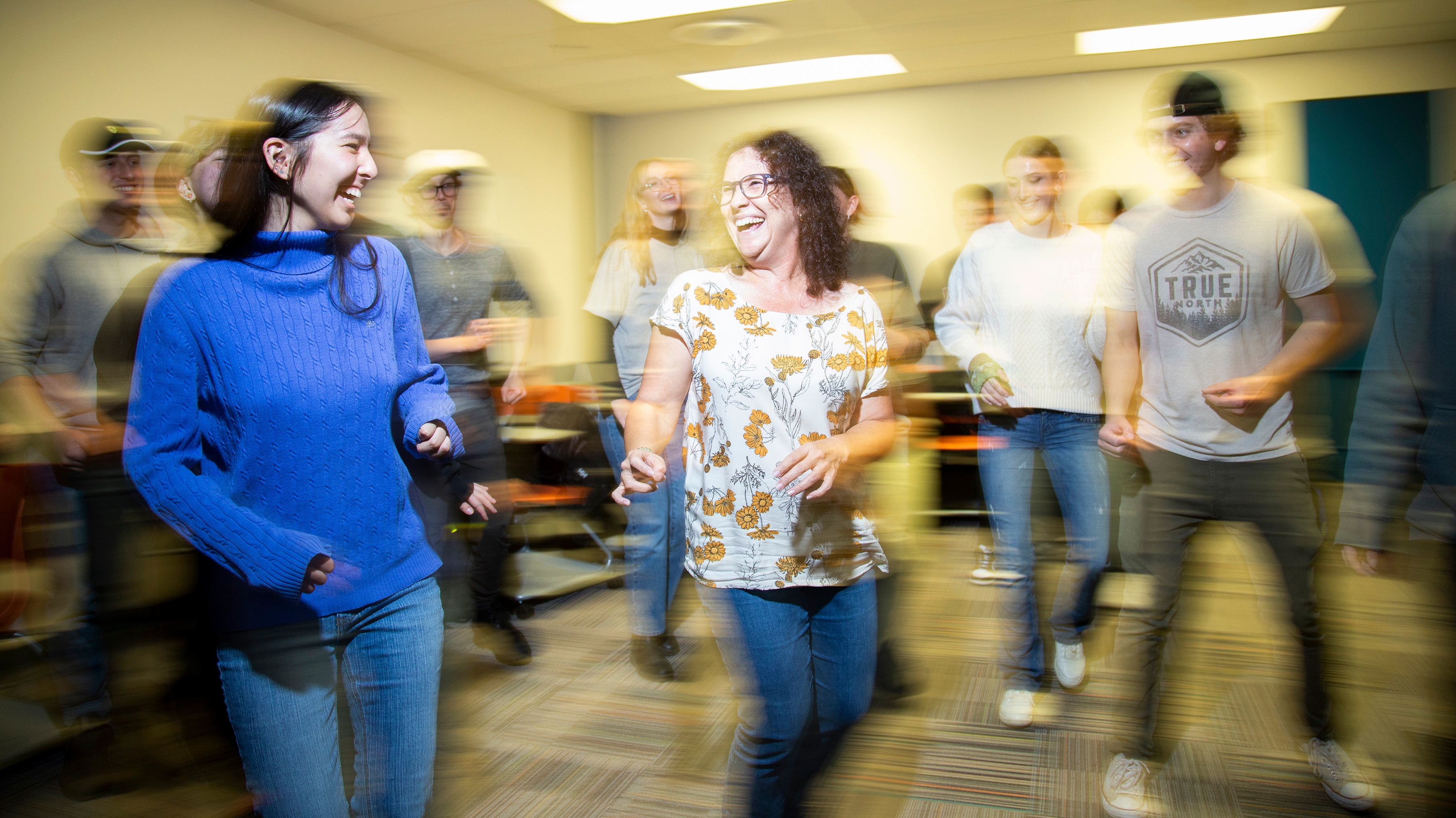 Alicia Ulysses, a professor with curly long hair and glasses, teaches Spanish through dance. Around her, students in her class are dancing and smiling. The photo captures the movement of students through slight blurring and light effects.