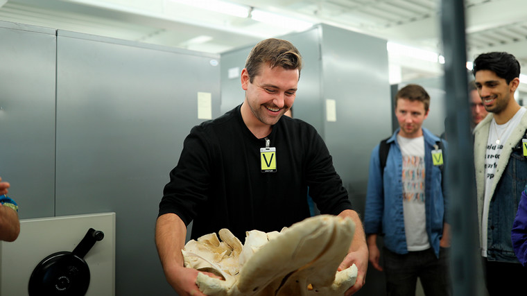 A student is grinning as he holds a large piece of whale bones in the archives of the Royal BC Museum. Other students are watching him in the background.