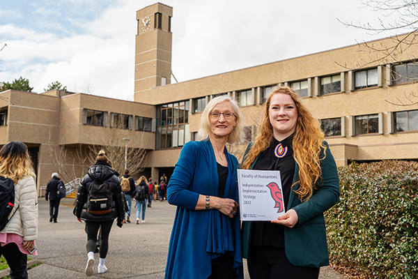 Lisa Surridge and Lydia Toorenburgh standing in front of the Clearihue building, holding a copy of the new Indigenization Implementation Strategy 
