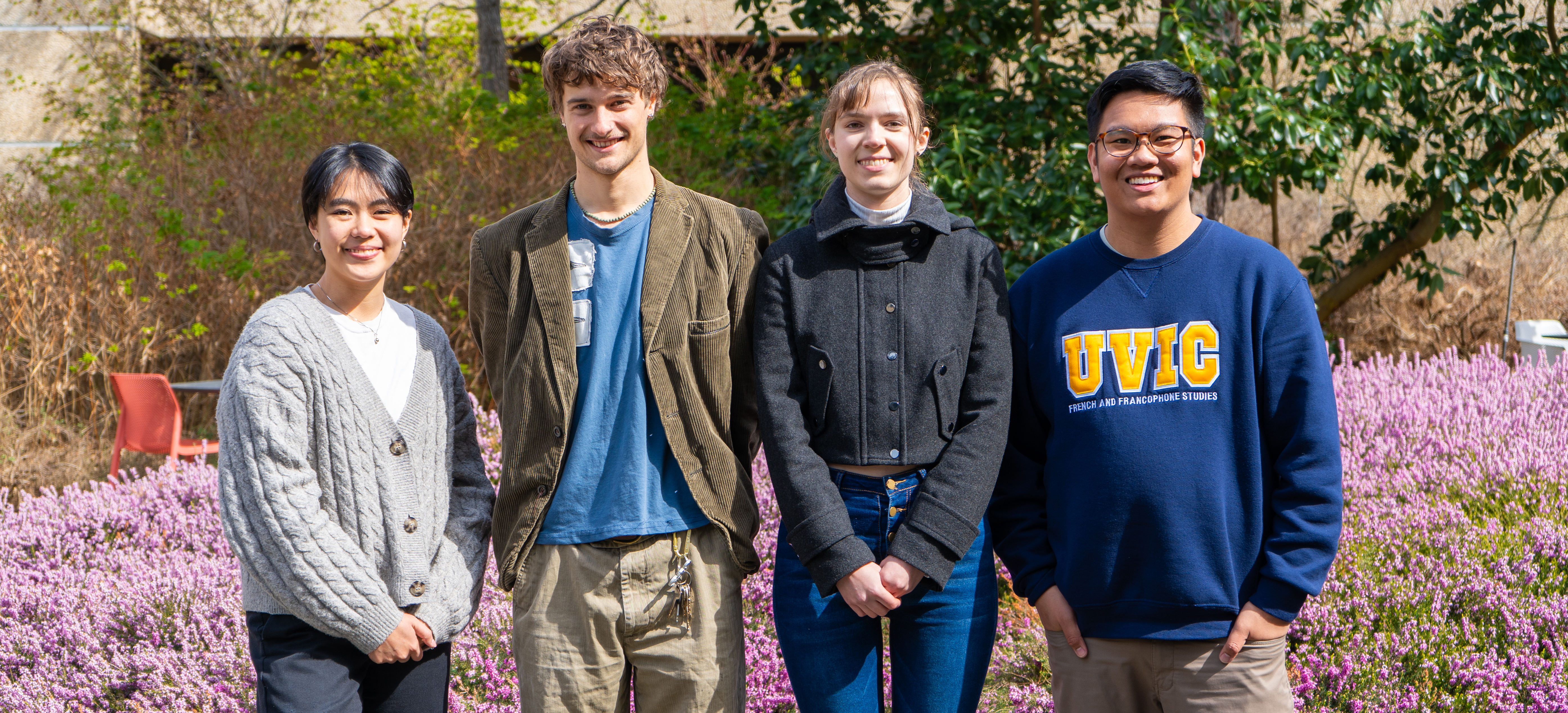 Four students are smiling on a sunny day in the Clearihue Courtyard, posing in front of blooming purple flowers on shrubs.
