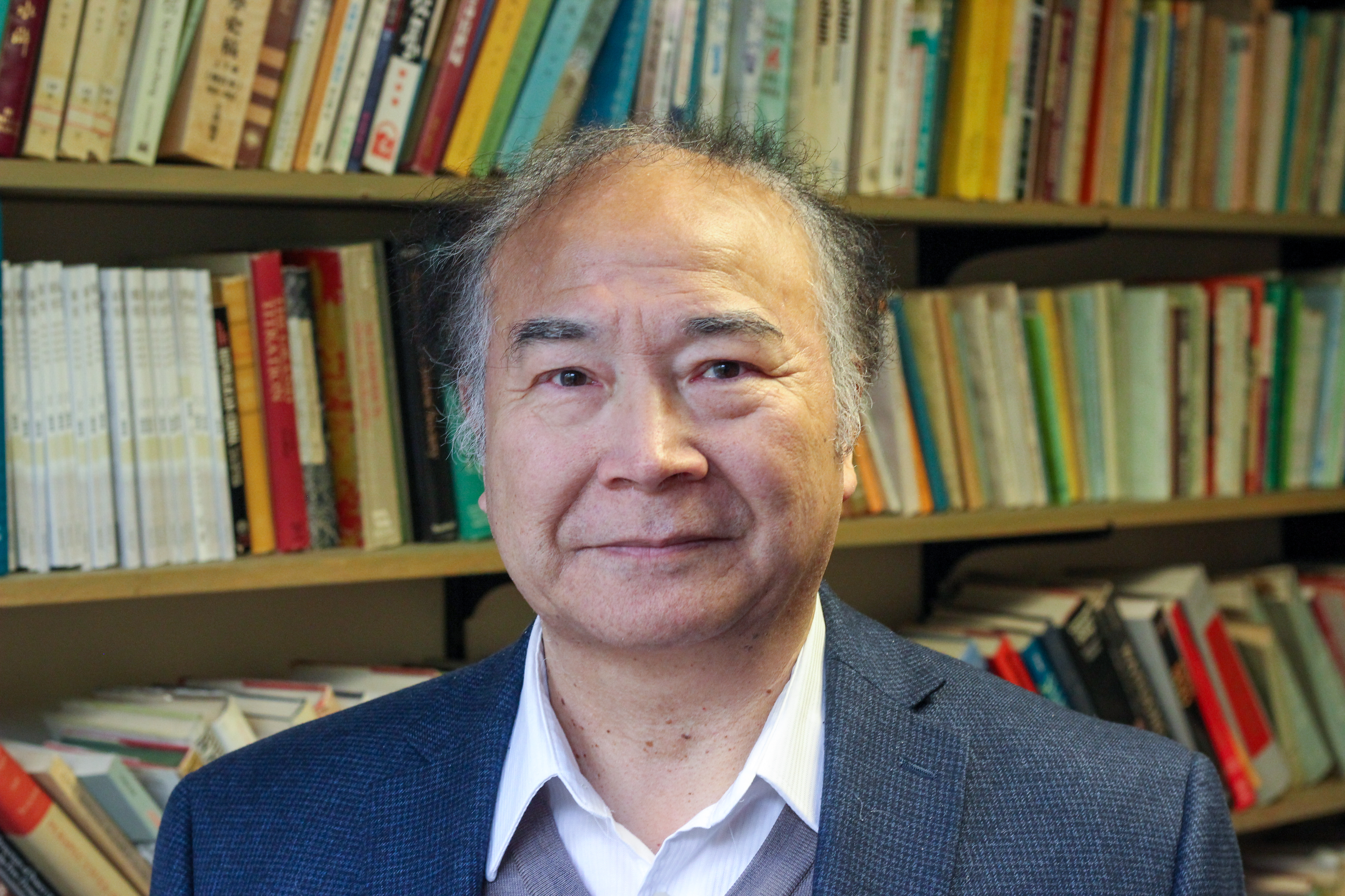 A headshot of Zhongping Chen, a Chinese man with grey-and-black hair wearing a blue blazer, grey sweater and white collared shirt, in front of a full bookcase.