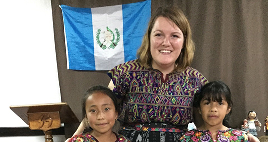 Rebecca Oudman standing with two girls in front of a Guatemala flag