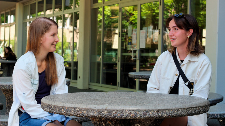 Two French students are sitting at a round picnic table outside the UVic Library, talking.