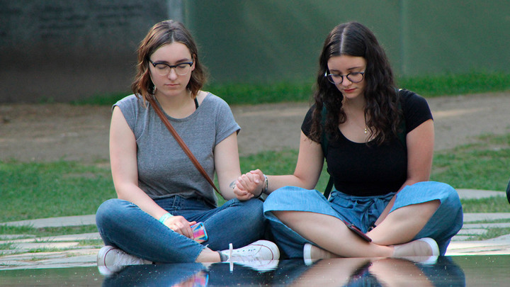 Two students are sitting solemnly holding hands at a memorial site, in front of a reflective pool.