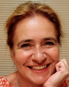 A headshot profile photo of Joanna Schaller smiling at the camera with one hand resting under her chin. She has light brown hair, brown eyes, and is seated indoors before a plain backdrop of closed white window blinds. 