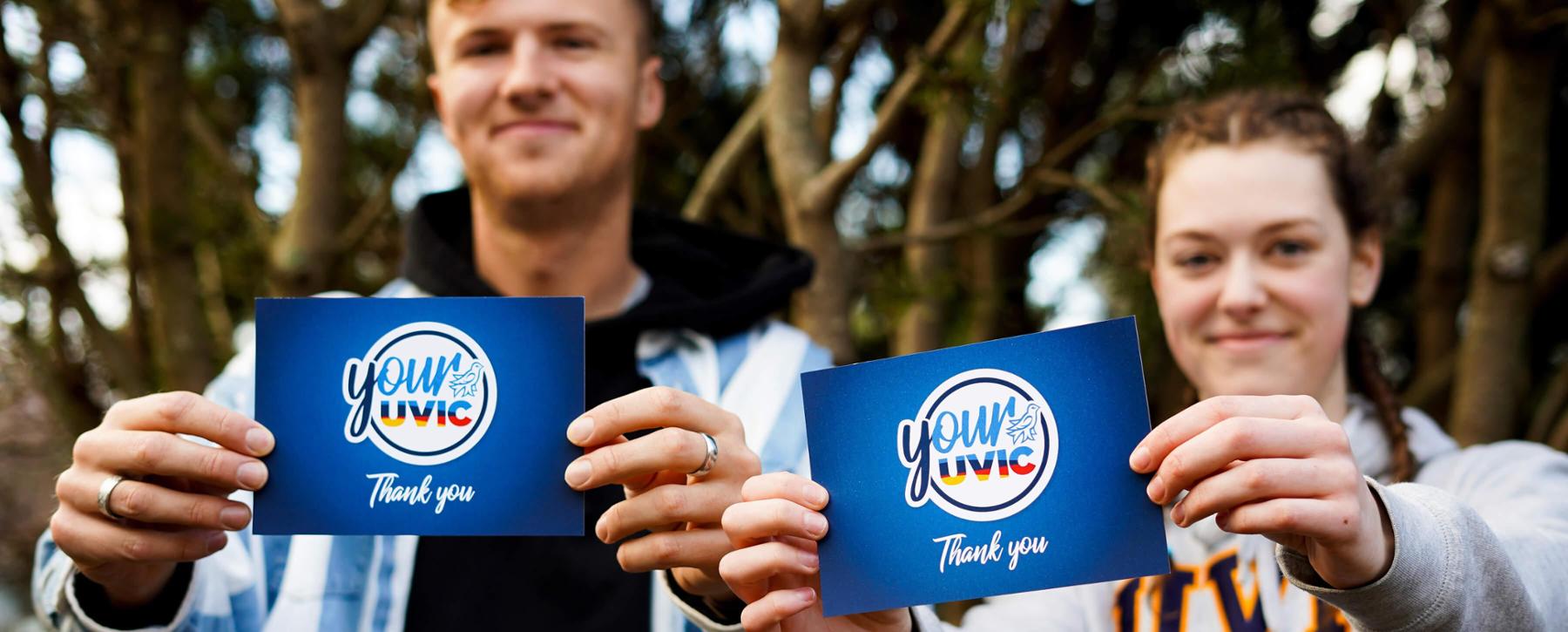 Two people stand smiling at the camera holding blue cards that say thank you with the Your UVic logo