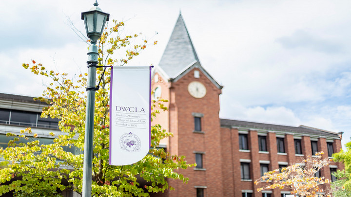 A banner hanging from a lamppost reads 'DWCLA – Doshisha Women’s College of Liberal Arts, Founded in 1876.' In the background, there is a red-brick building with a clock tower and arched windows. Trees with green and yellow leaves frame the scene under a partly cloudy sky
