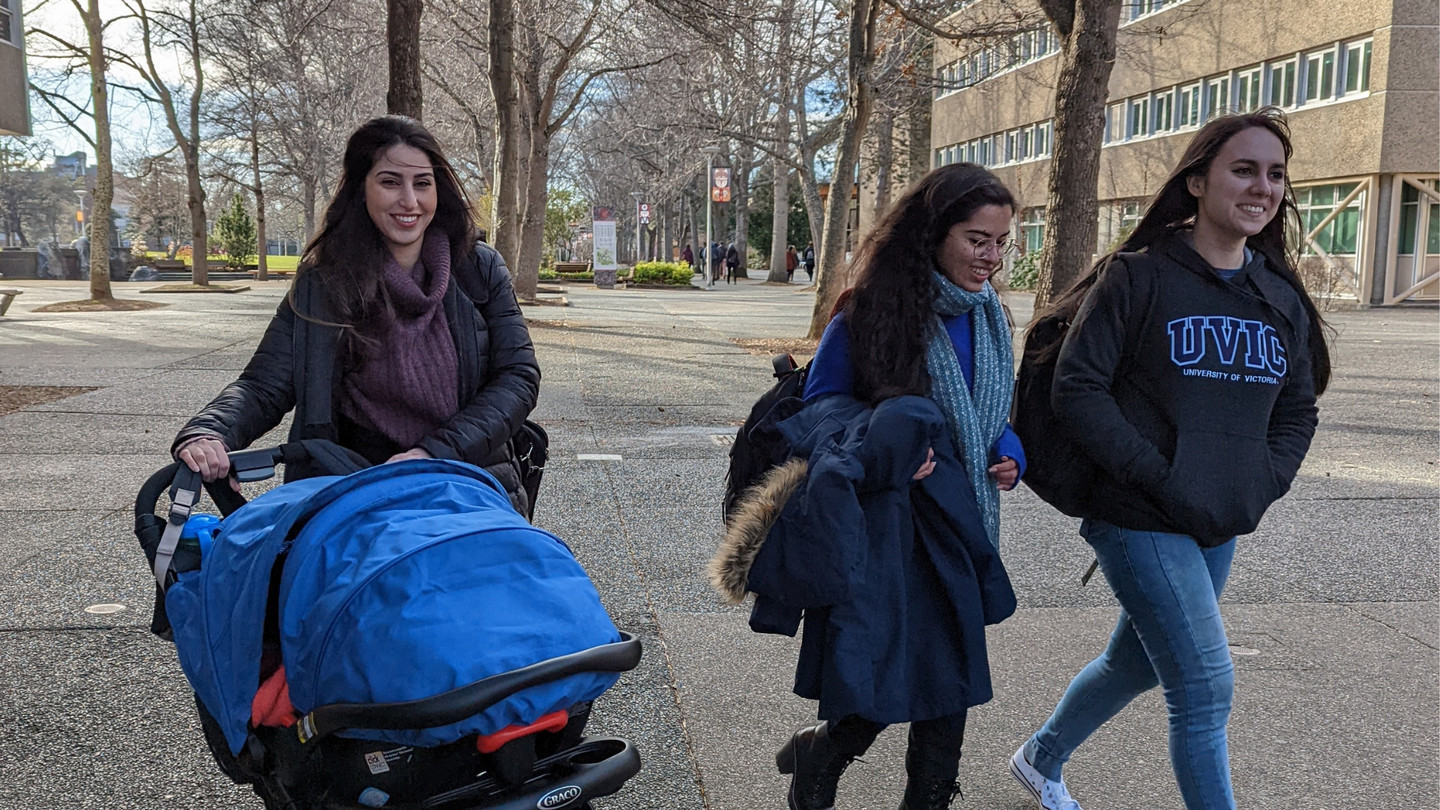 3 students walking through the quad at UVic, one is pushing a stroller