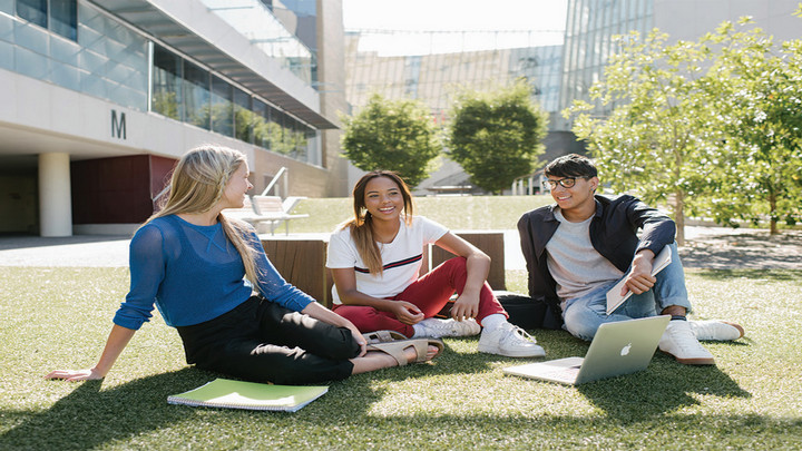 Three students sit around a laptop and notebook on the grass in front of Victoria University lecture halls.