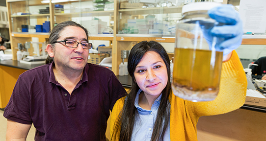 Juanes and Olson in a lab looking at a jar containing brownish contents