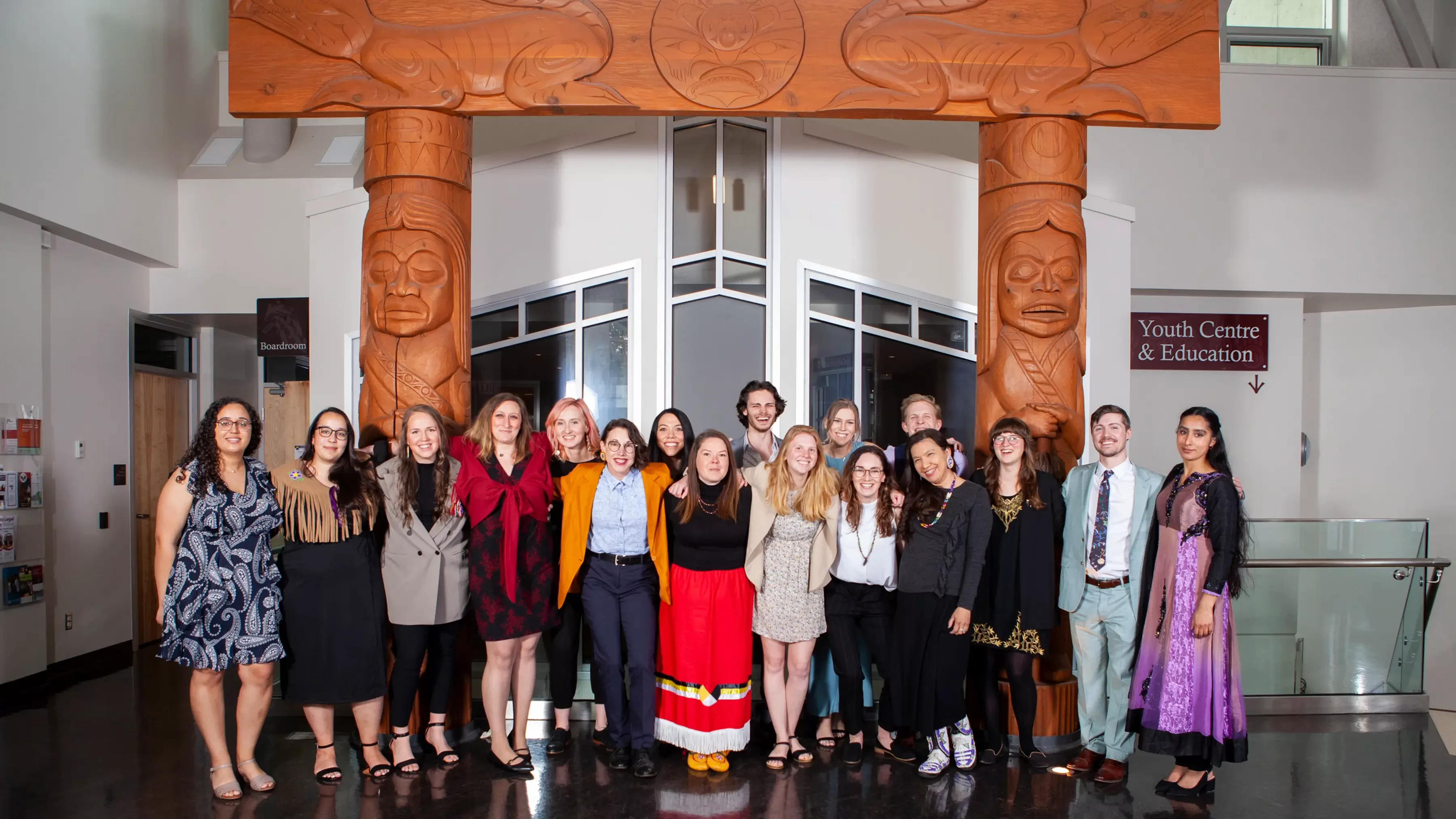 A row of graduating students stand as a group in front of Indigenous woodwork.