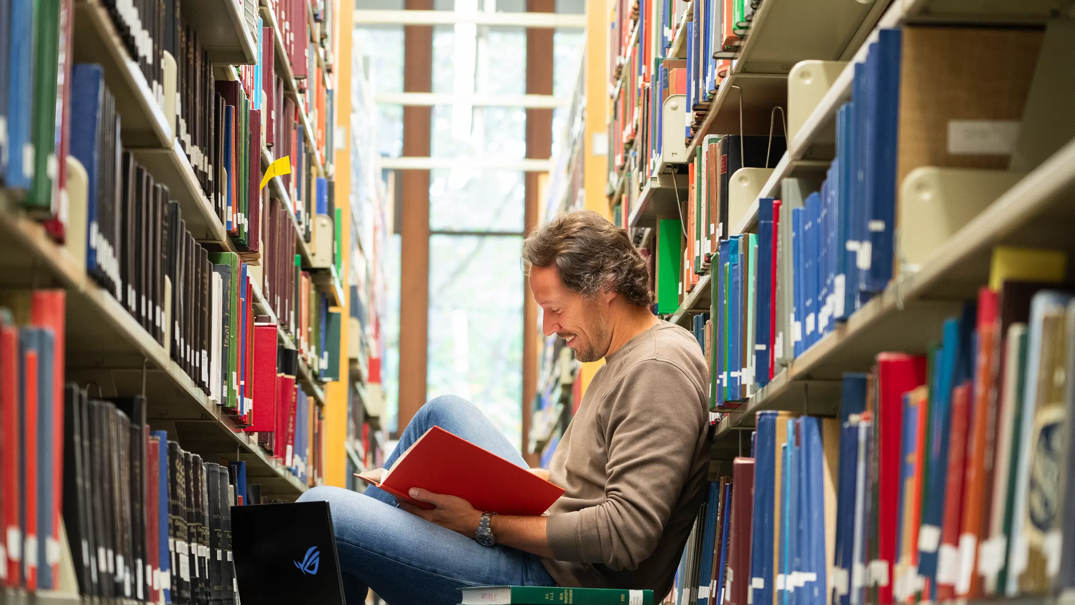 A man sits reading in between 2 rows of librray bookshelves. 